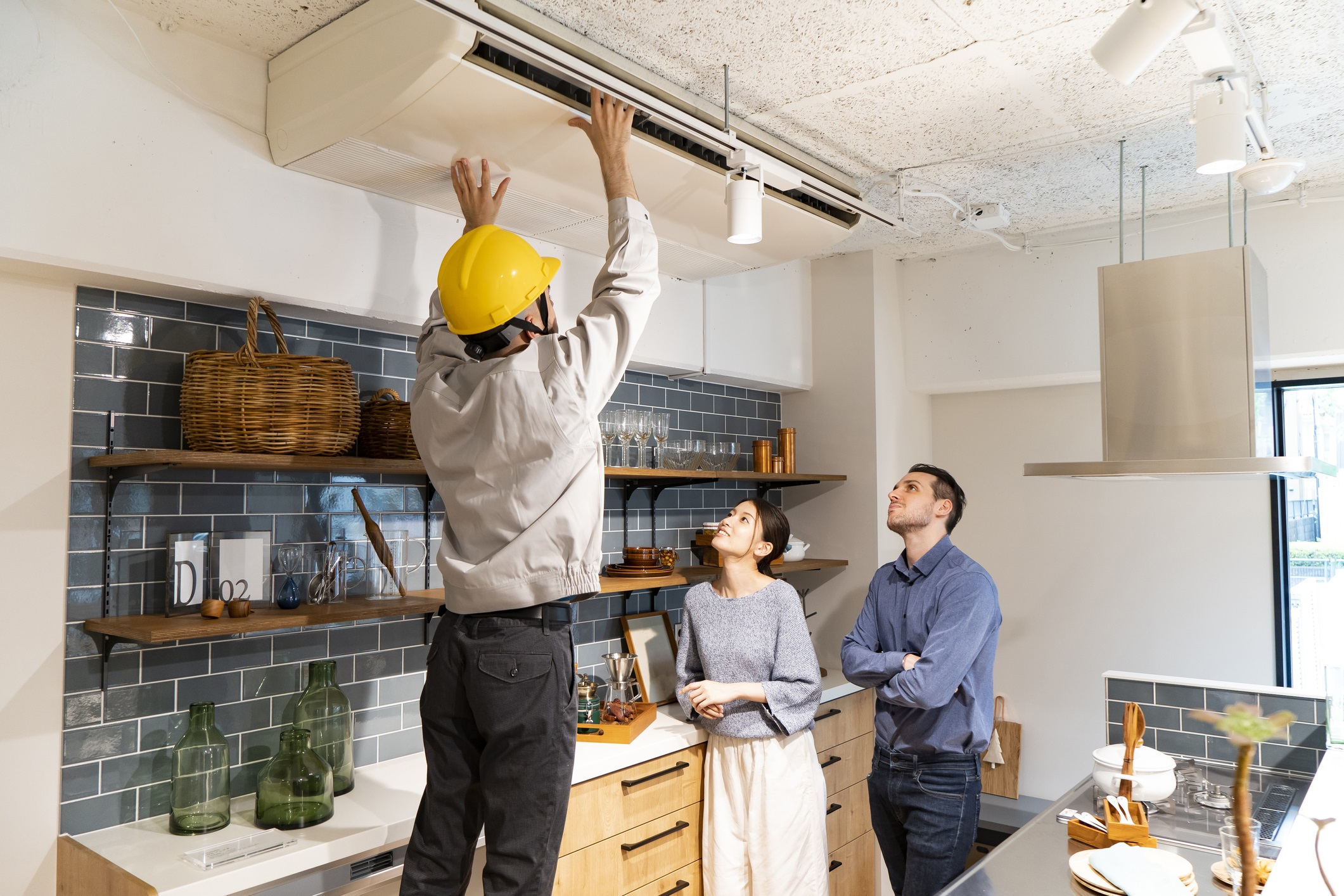 A repairman wearing a yellow helmet checks an air conditioner as a male and female couple watches in their apartment's kitchen for residents wish list blog.